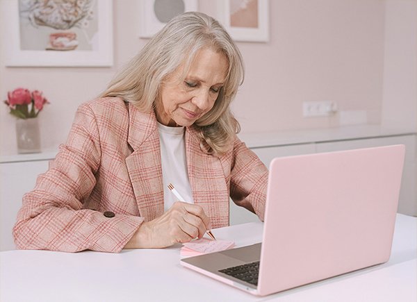 Senior woman writing notes while working on a laptop, symbolizing reinvention, purpose, and new beginnings.